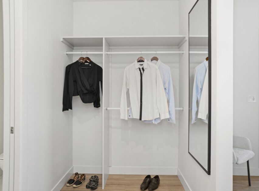 A minimalist walk-in closet with white shelves holding several shirts and a black blouse on hangers, shoes on the floor, and a large wall mirror on the right. The space is bright with light-colored flooring.