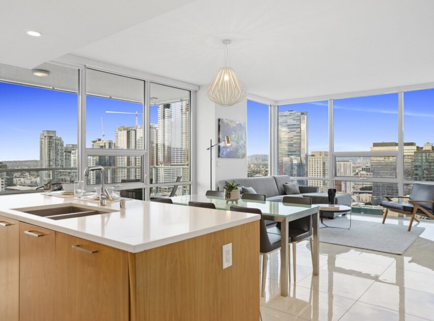 Modern high-rise apartment with floor-to-ceiling windows, city skyline views, an open kitchen with a center island, dining area, and a living room with a gray sofa and contemporary decor. Bright and spacious atmosphere.