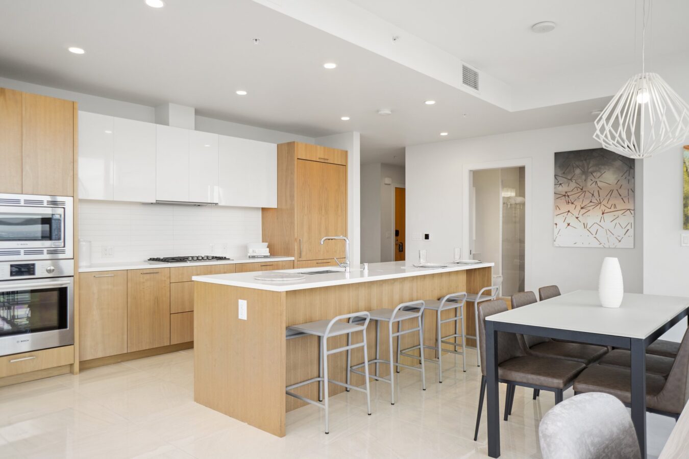 Modern kitchen with light wood cabinets, white countertops, built-in stainless steel appliances, a large island with four gray stools, and an adjacent dining table with six chairs. Minimalist decor and bright lighting.