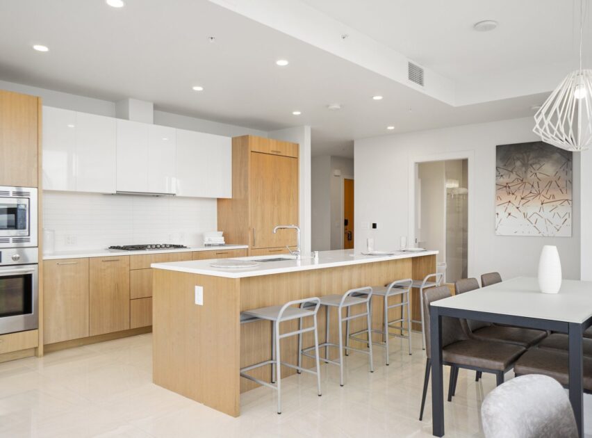 Modern kitchen with light wood cabinets, white countertops, a central island with four gray stools, stainless steel appliances, a dining table with six chairs, and minimalist decor in a bright, open space.