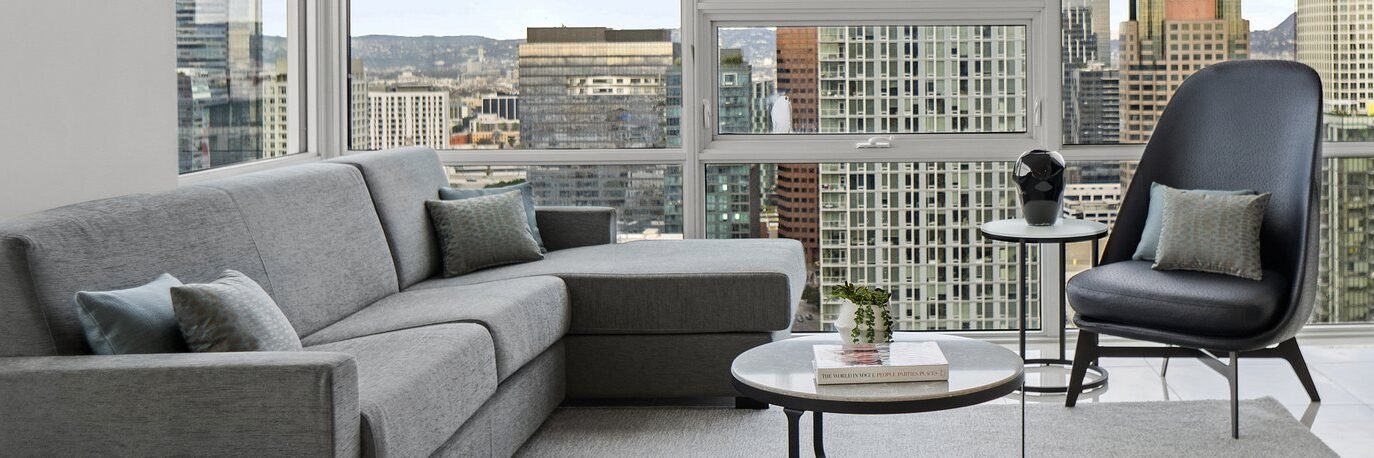 Modern living room with a grey sectional sofa, accent pillows, a black lounge chair, round coffee table with books and a small plant, large windows offering a city skyline view with tall buildings.