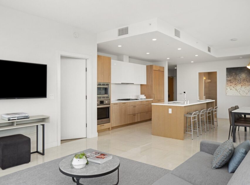 Modern open-concept living space with a sleek kitchen featuring wood cabinetry, an island with four stools, and stainless steel appliances. A living area with a gray sofa and coffee table faces a wall-mounted TV.