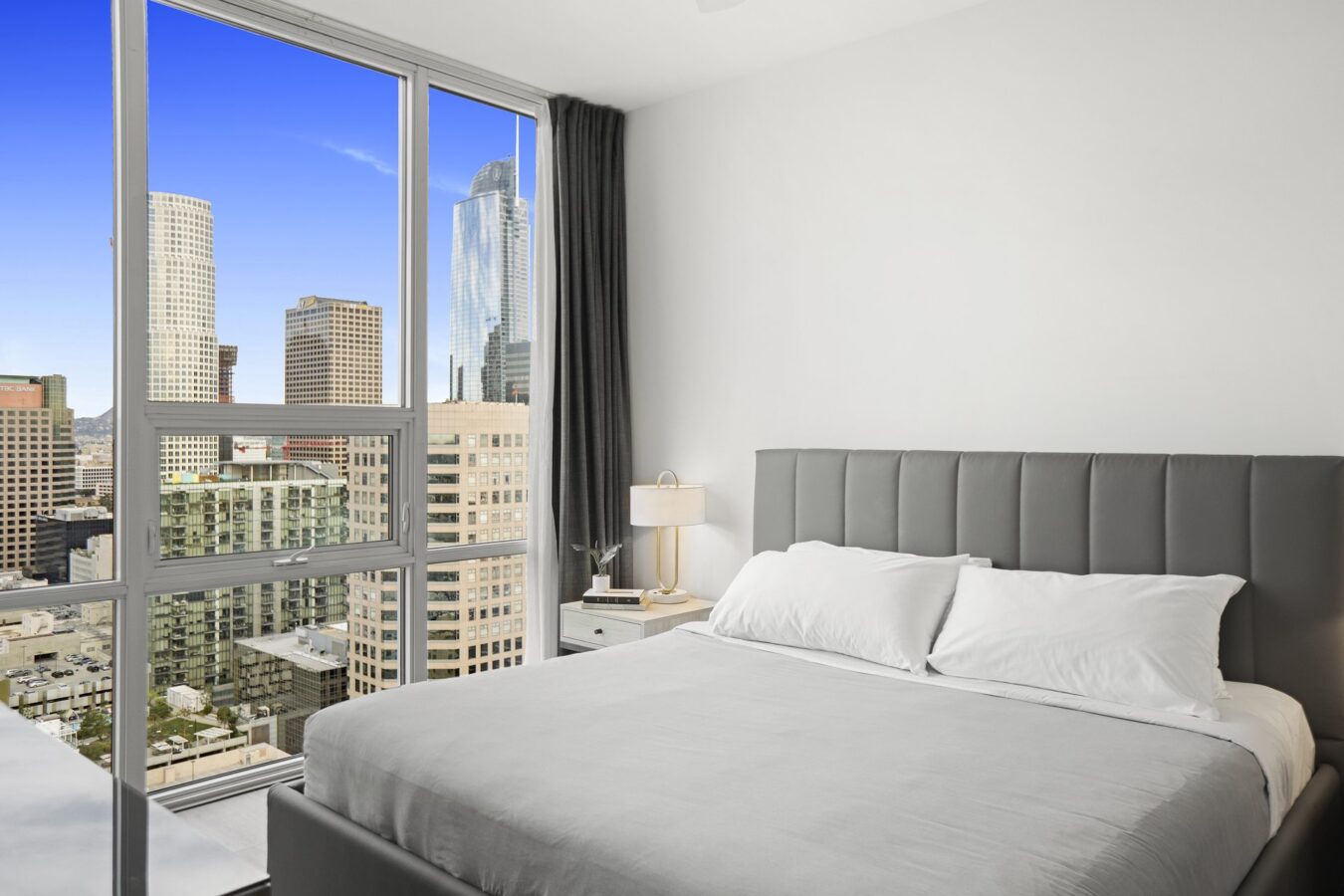 Modern bedroom with a neatly made gray bed, white walls, and large floor-to-ceiling windows offering a view of downtown city skyscrapers under a clear blue sky. A nightstand with a lamp sits by the bed.
