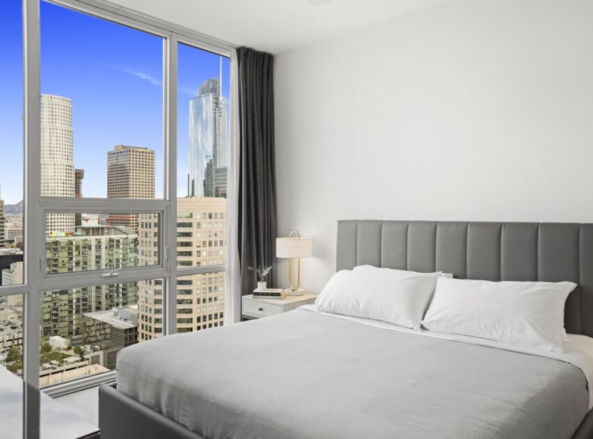 Modern bedroom with a large bed, gray headboard, and gray bedding. Floor-to-ceiling windows reveal a city skyline with tall buildings and a clear blue sky. A nightstand with a lamp sits beside the bed.