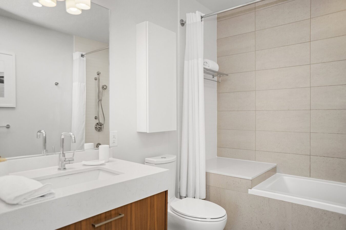 Modern bathroom with a white countertop sink, wooden vanity, wall-mounted toilet, and a bathtub with beige tile surround and white shower curtain. A mirror and towel are visible, creating a clean, minimalist look.