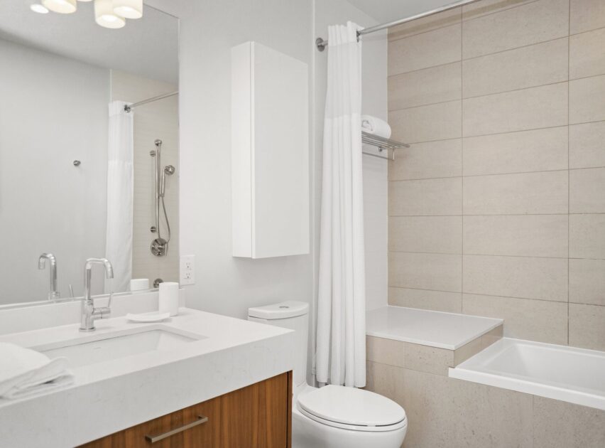 Modern bathroom with a white sink, wooden vanity, toilet, and bathtub with a white shower curtain. Neutral tiled walls and a mounted towel above the tub create a clean, minimalist look.