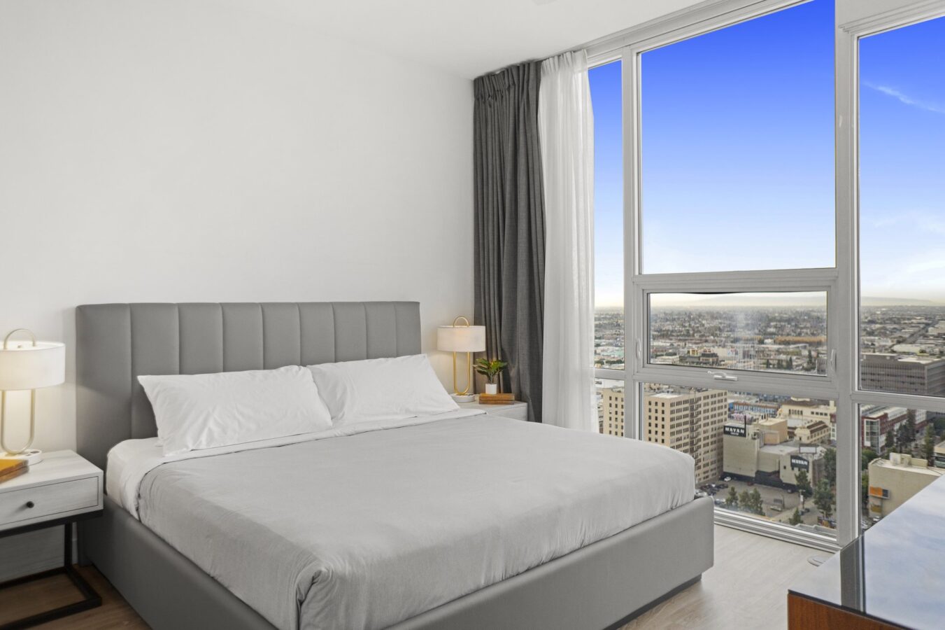 Modern bedroom with a large gray bed, white walls, two bedside tables with lamps, and a big window showing a city view under a clear blue sky.