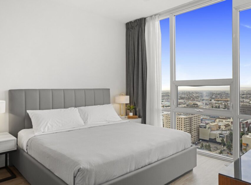 A modern bedroom with a gray upholstered bed, white bedding, nightstands, and a large window offering a cityscape view under a clear blue sky.