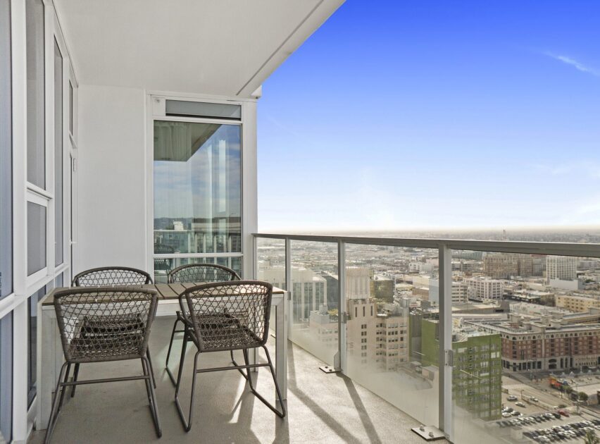 A modern high-rise balcony with a glass railing, a table, and four chairs, overlooking a cityscape under a clear blue sky.