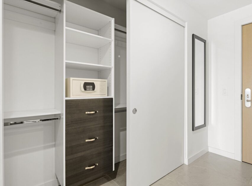 A modern closet with open shelves and drawers, featuring a digital safe on the top shelf. There is a sliding white door, a large wall mirror, and a wooden entry door visible. The space is clean and minimalistic.