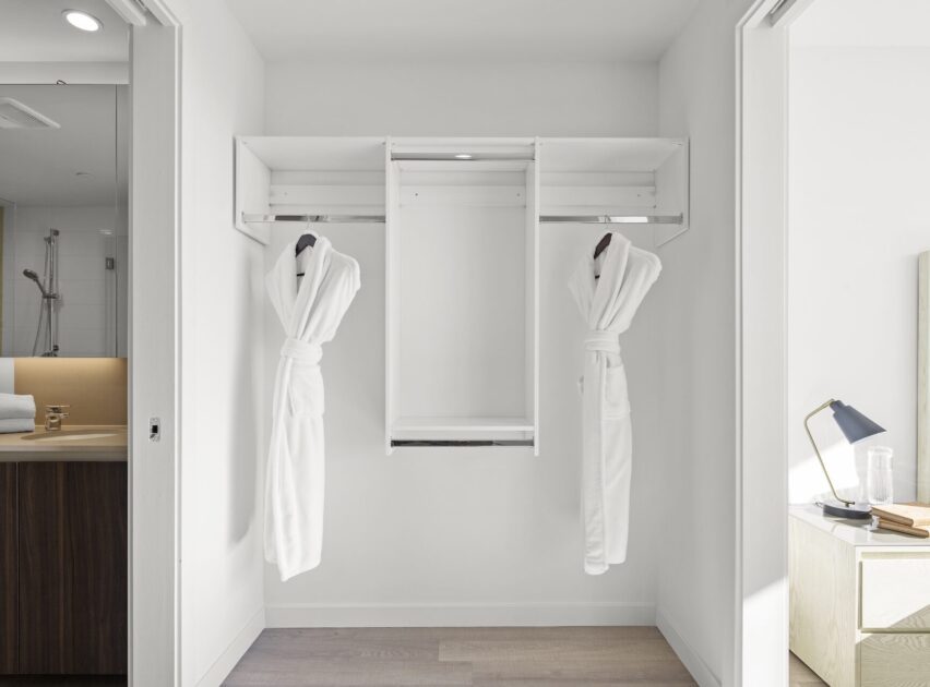Minimalist walk-in closet with two white bathrobes hanging on either side of a central shelf. The area is bright and modern, with an adjacent bathroom on the left and a bedside table with a lamp on the right.