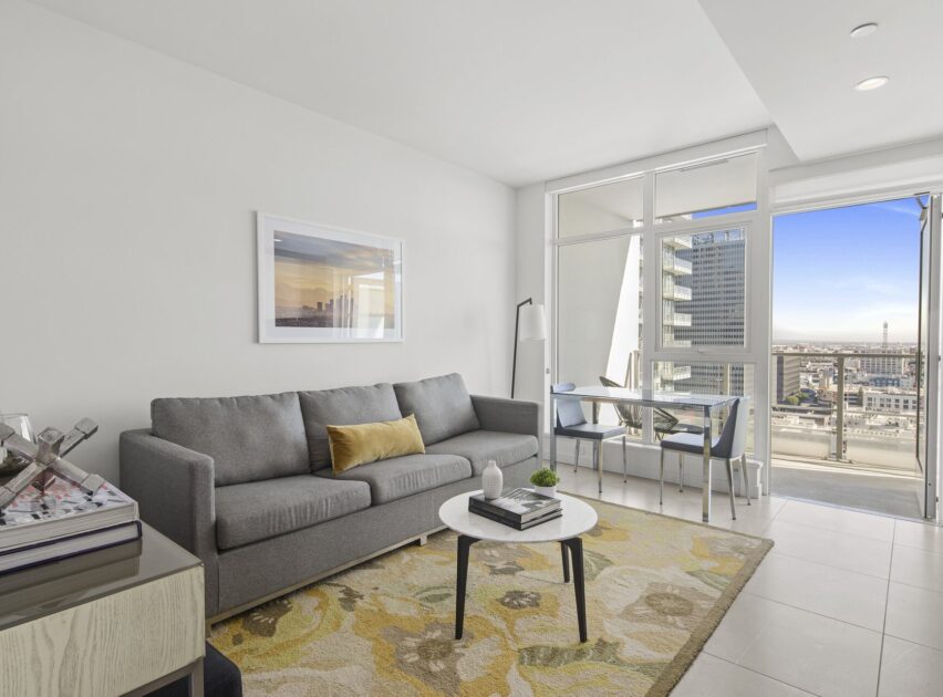 Modern living room with a gray sofa, yellow-accented rug, round coffee table, and a small dining area by large windows showing a city view and access to a balcony under a clear sky.