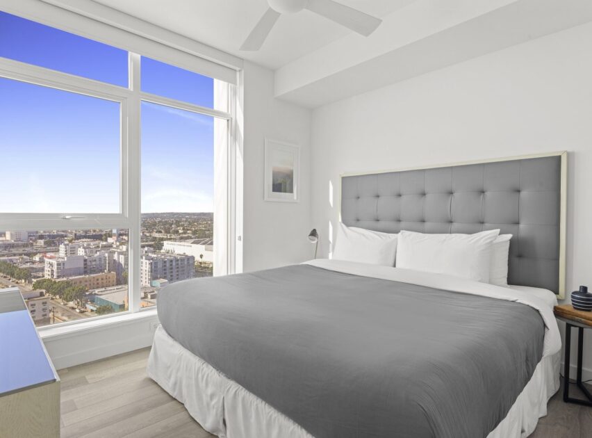A bright, modern bedroom with a large window showing a city view, a neatly made bed with a gray comforter and white pillows, light wood floors, and minimal decor.