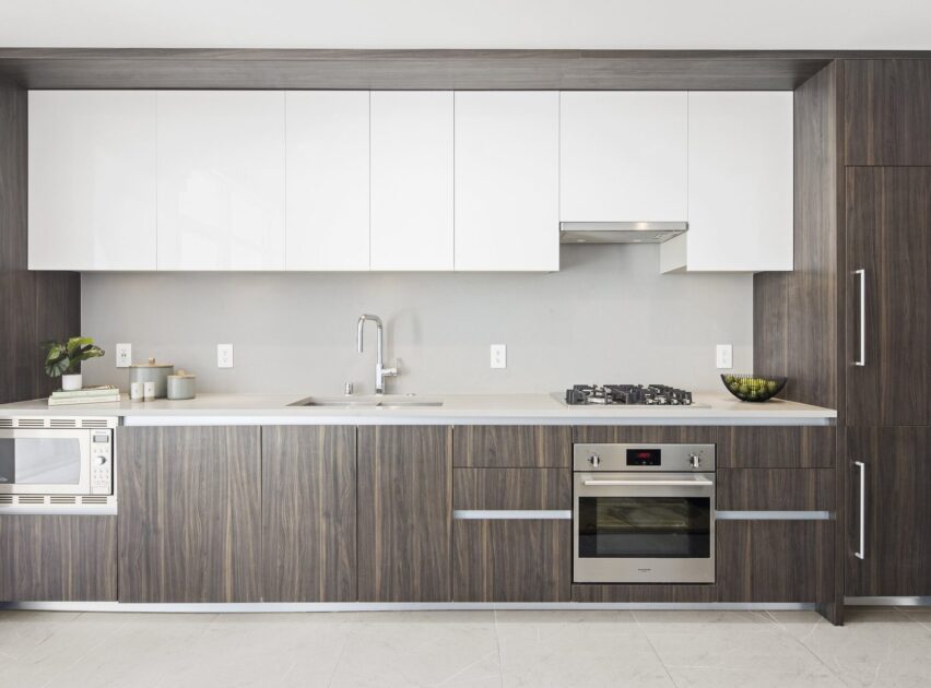Modern kitchen with wood cabinets, white upper cabinets, built-in oven, gas cooktop, microwave, sink, and minimalist decor. The countertop is white, and the backsplash is light gray. The space is clean and uncluttered.