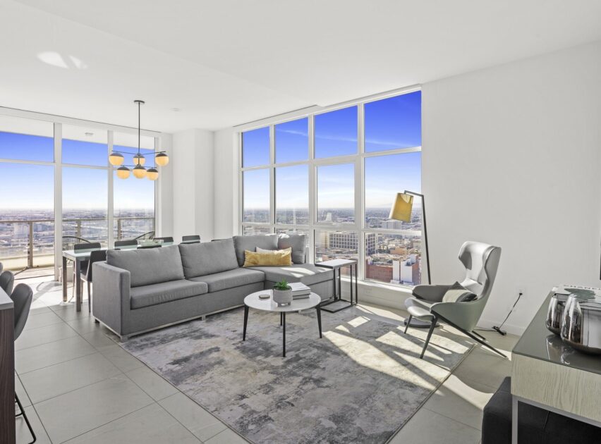 Bright, modern living room with large windows showing a cityscape, gray sofa, armchair, floor lamp, coffee tables, rug, dining area, and light fixture, all in a minimalist, open-plan design.