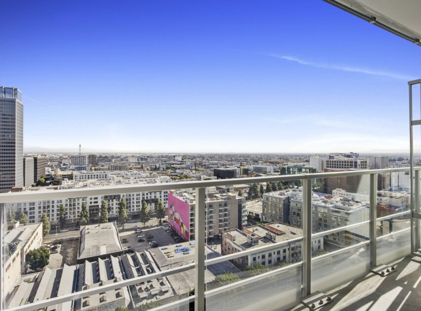 View from a high-rise balcony overlooking a cityscape with modern buildings, a colorful mural, and clear blue sky above. The glass railing runs along the edge of the balcony.