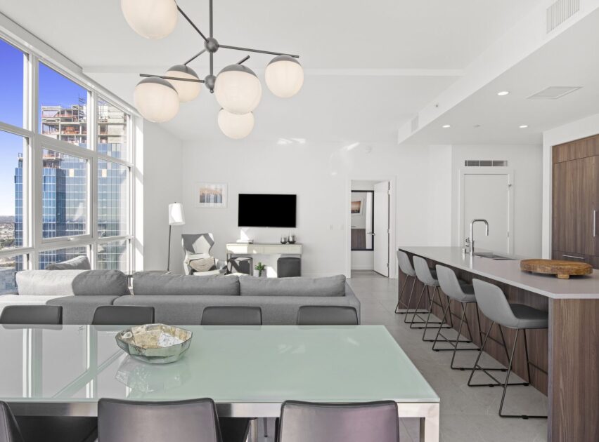 Modern open-concept living space with large windows, gray sectional sofa, glass dining table, sleek kitchen island with barstools, and a minimalist decor. City buildings visible outside.
