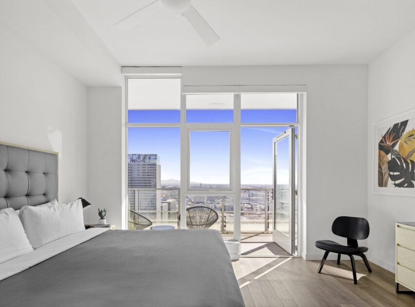 A modern, bright bedroom with a large bed, minimalist furniture, and floor-to-ceiling windows leading to a balcony with city views under a clear blue sky.