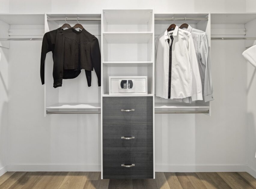 A modern walk-in closet with hanging black and white shirts, a black jacket, a small safe on a shelf, and three drawers below, all set against white walls and wood flooring.