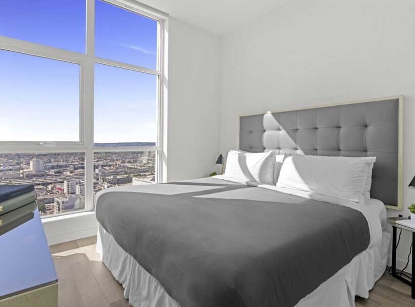 Bright bedroom with large windows overlooking a cityscape, featuring a gray upholstered headboard, neatly made bed with white and gray bedding, and modern minimalist decor.