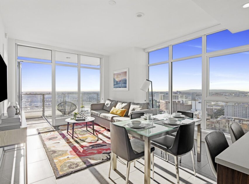 Bright modern living room with large windows, a gray sofa, colorful rug, glass dining table set for four, and a city skyline view. Natural light fills the space, creating an open and airy atmosphere.