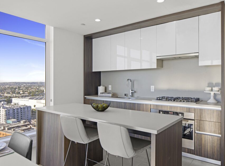 Modern kitchen with sleek white and wood cabinetry, a built-in oven, a gas cooktop, and a large window with a city view. A kitchen island with three gray chairs and a bowl sits in the center.