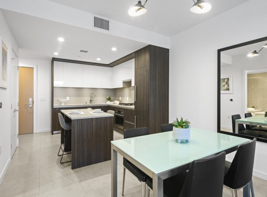 Modern kitchen and dining area with white cabinets, dark wood accents, a kitchen island set for two, a glass dining table with four black chairs, a large wall mirror, and a potted plant centerpiece.