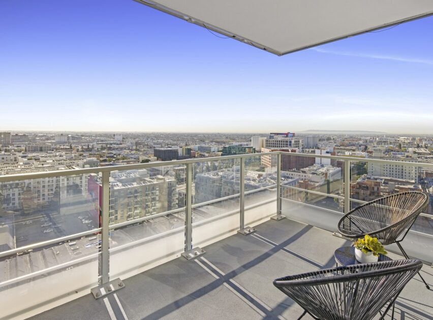 A modern balcony with glass railing overlooks a cityscape under a clear blue sky; two black chairs and a small table with a plant are arranged near the edge.