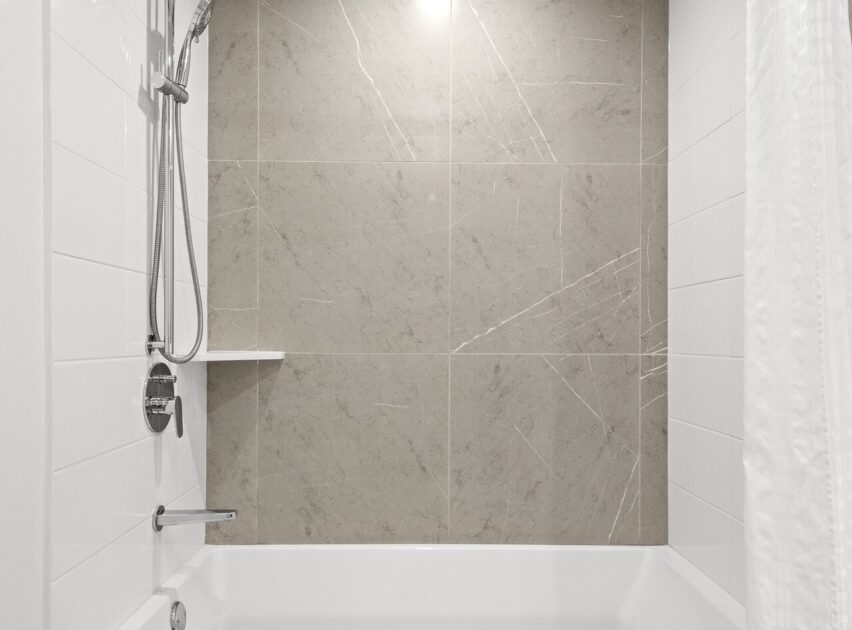 A modern bathtub with a wall-mounted showerhead, white tile walls, a gray tiled accent wall with subtle veining, and a small corner shelf. A white shower curtain is partially visible on the right.