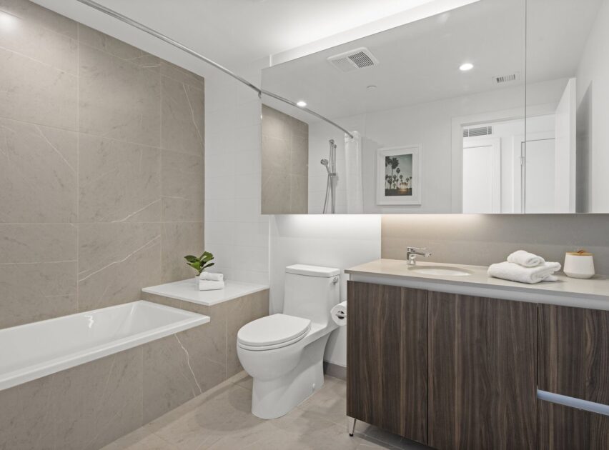 Modern bathroom with a bathtub, toilet, wood vanity with a sink, large mirror, and light gray tiled walls. A small plant, towels, and soap dispenser sit on the countertop; framed art hangs on the wall.