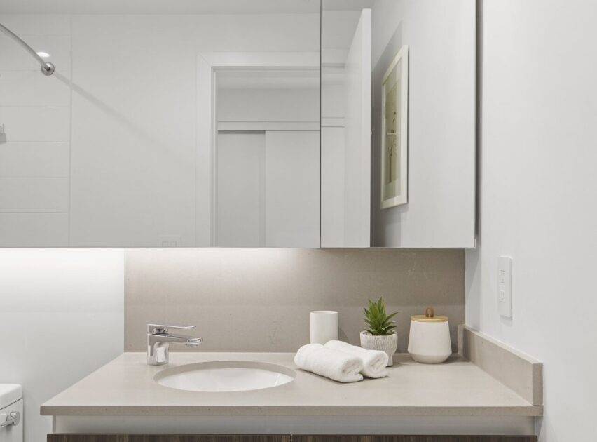 Modern bathroom with a light-colored countertop, a sink, a silver faucet, rolled white towels, a small potted plant, and decorative containers. A mirror and framed picture are on the white wall above the sink.