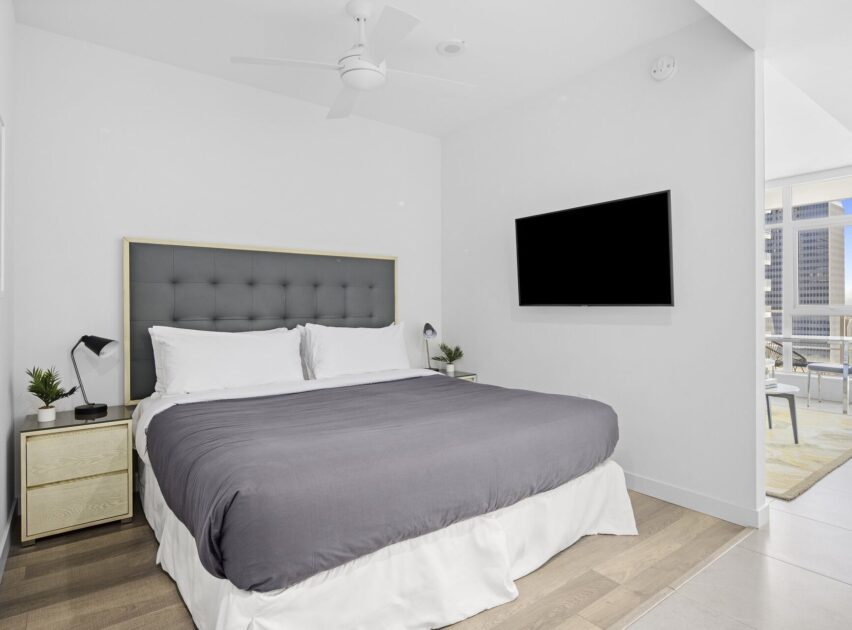 Modern bedroom with a king-sized bed, gray headboard, gray comforter, and white walls. There are two nightstands with small plants, a wall-mounted TV, ceiling fan, and large window with city view in the adjacent room.