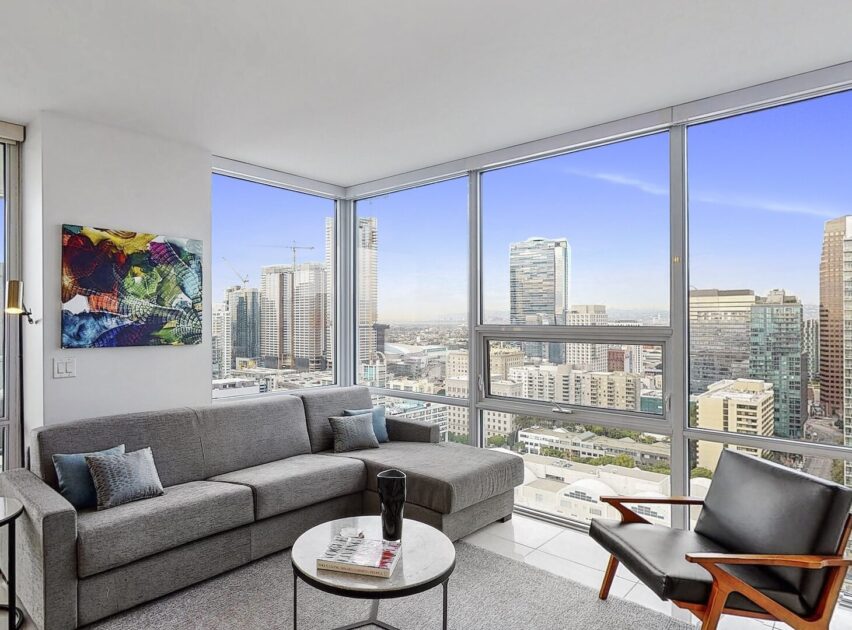 A modern living room with a gray sectional sofa, an armchair, a small round coffee table, colorful wall art, and floor-to-ceiling windows offering a city skyline view on a clear day.