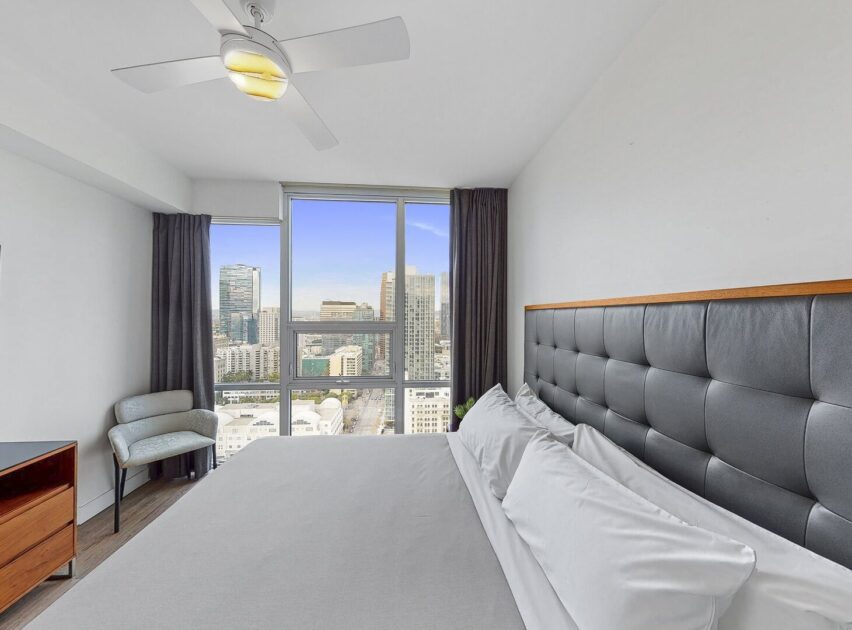 Modern bedroom with a large bed, white linens, a padded headboard, a ceiling fan, a small dresser, and a chair by floor-to-ceiling windows offering a city skyline view.
