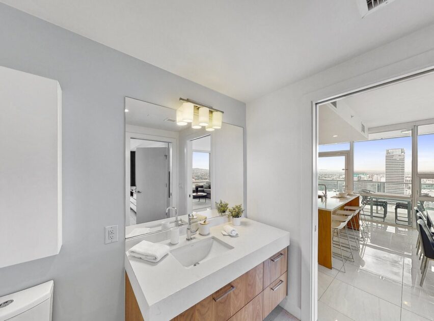 Modern bathroom with a large mirror, rectangular sink, and wooden vanity. Through the open door, a bright kitchen and dining area with large windows and city views are visible.