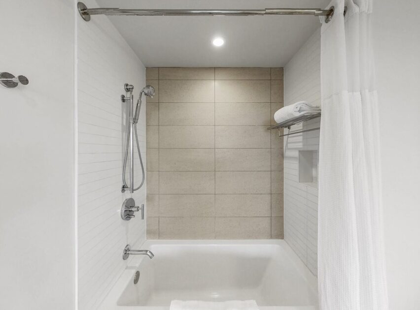 A modern bathtub and shower with a beige tiled back wall, white side walls, a mounted showerhead, a towel rack with folded towels, and a white shower curtain partially open.