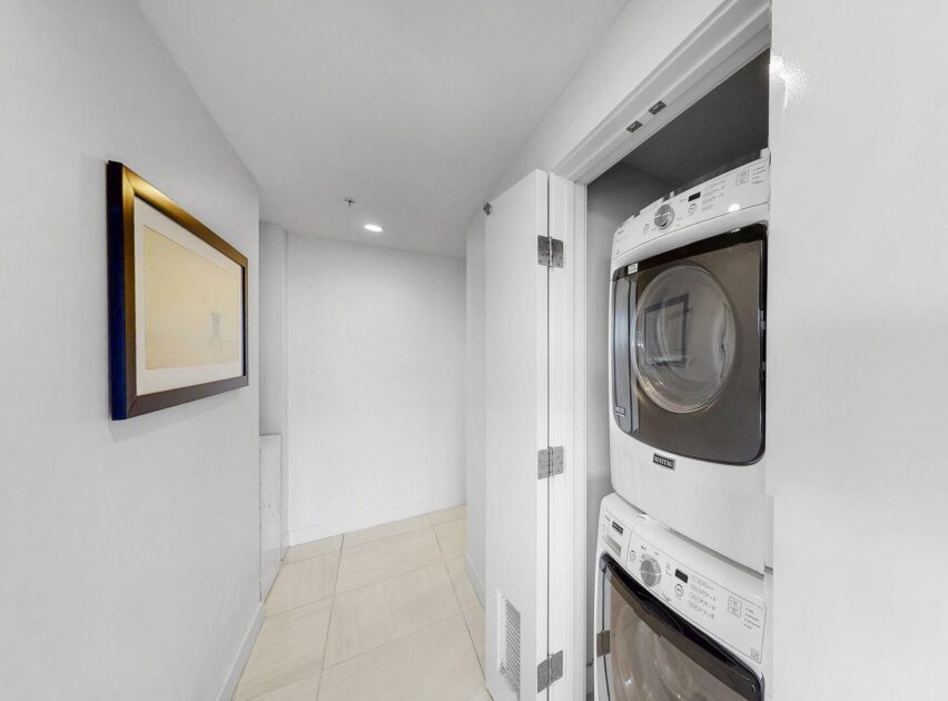 A small laundry area with a stacked white washer and dryer, partially enclosed behind double doors. The space has light-colored tile flooring, white walls, and a framed picture hanging on the left wall.