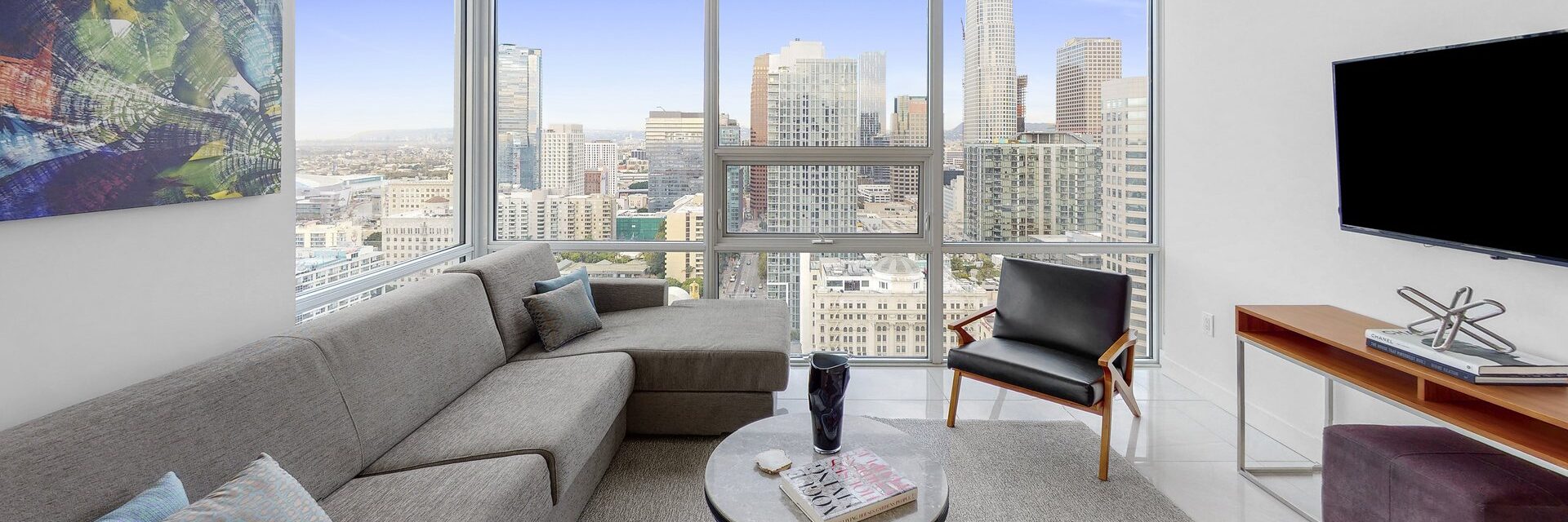 A modern living room with a gray sectional sofa, black armchair, round coffee table, and wall-mounted TV. Large windows reveal a city skyline with tall buildings and blue sky. Abstract art hangs on the wall.