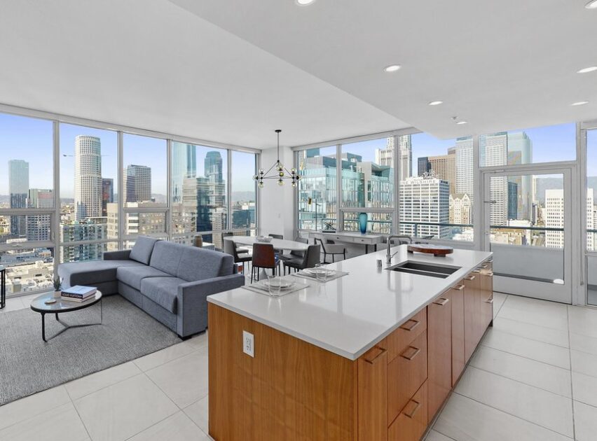 Modern high-rise apartment with floor-to-ceiling windows, a spacious open kitchen with an island, a gray sofa, dining table, and panoramic city skyline views in the background.