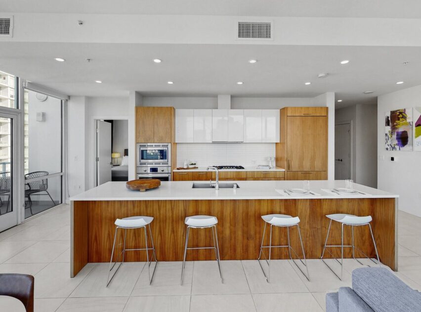 Modern kitchen with wood and white cabinetry, a large island with four white barstools, built-in appliances, and a dining area on a balcony visible through glass doors. Bright, open, and minimalistic design.