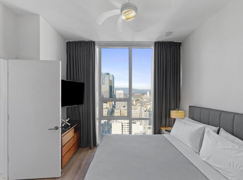 A modern, minimalist bedroom with a large bed, gray headboard, and gray curtains. A flat-screen TV is mounted near a dresser. Large windows offer a city skyline view under a clear sky.