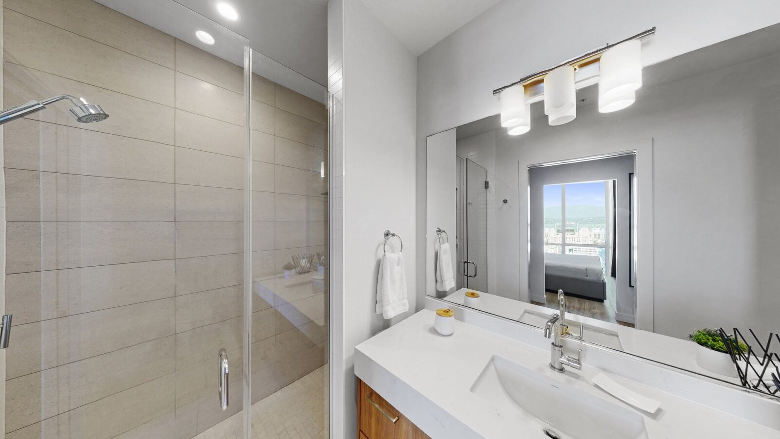 Modern bathroom with a glass-enclosed shower, white countertop with sink, towel ring, mirror, and overhead lights. An open doorway reveals a bedroom and large window with a city view.