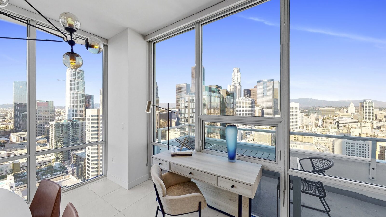 A modern apartment with large windows offering a scenic view of a city skyline. Inside, there’s a wooden desk with a blue vase, a chair, and a balcony with outdoor seating visible through the glass door.