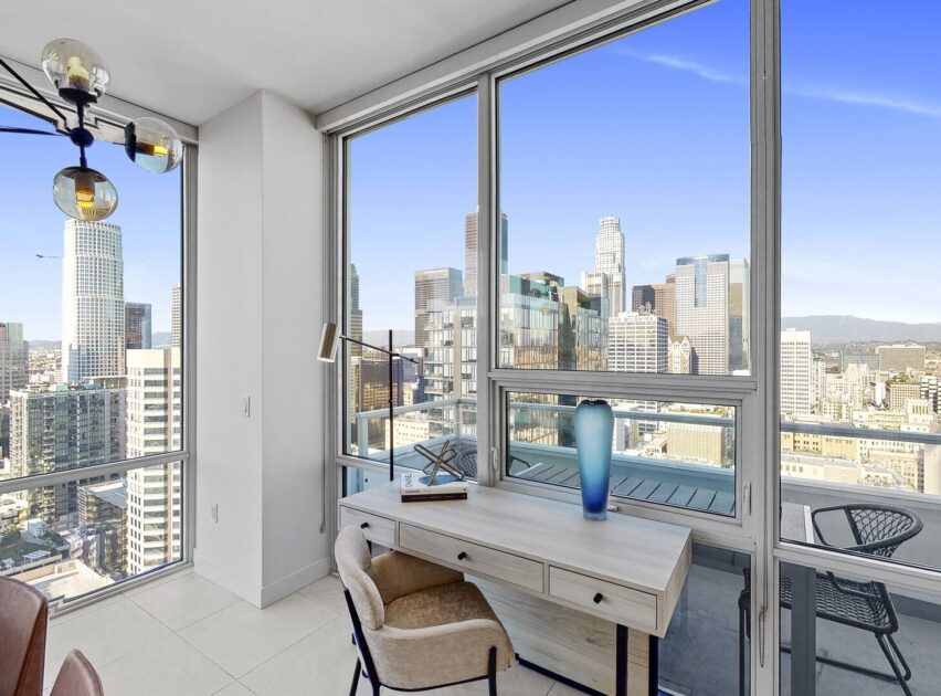 A modern apartment with floor-to-ceiling windows offers a view of a city skyline. Inside, there is a wooden desk with a blue vase, lamp, and chair next to a balcony with outdoor seating.