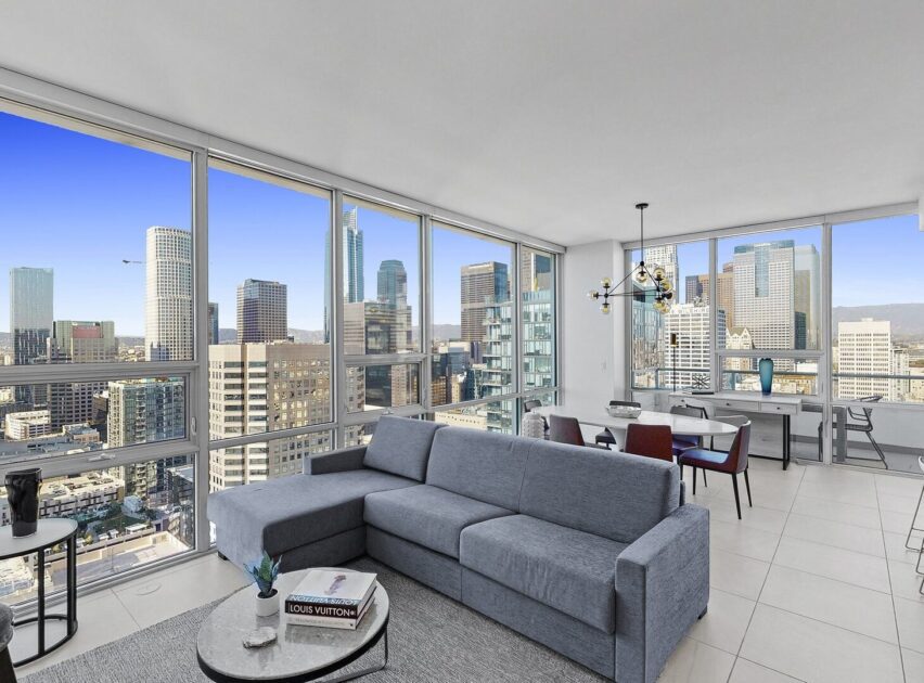 A modern high-rise apartment with large floor-to-ceiling windows offers a panoramic view of a city skyline. The room features a gray sectional sofa, a coffee table, and a dining area with a chandelier.