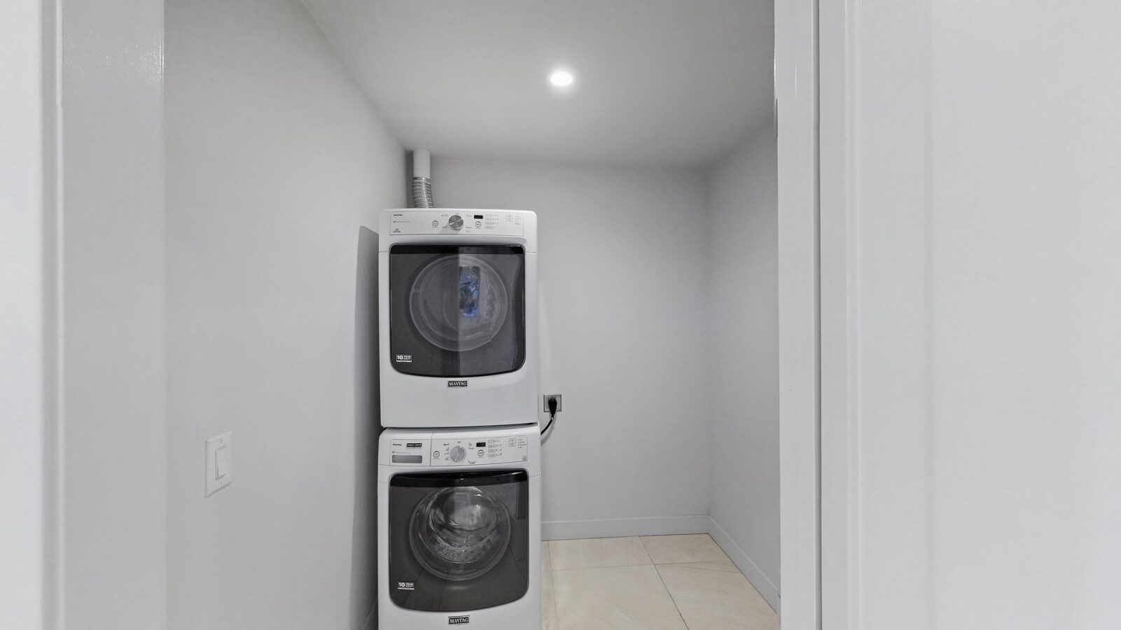 A small laundry room with white walls and floor tiles, featuring a stacked washer and dryer unit under a ceiling light.