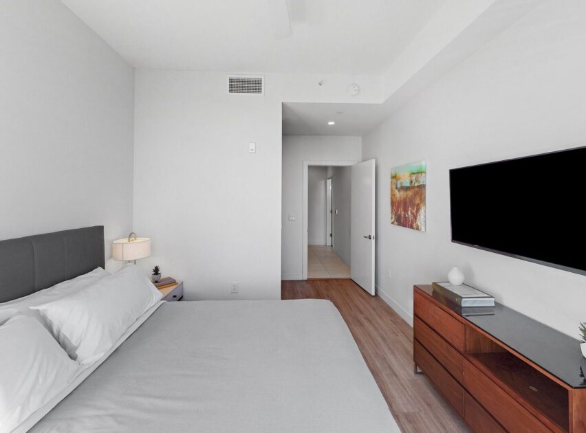 Minimalist bedroom with a large bed, white bedding, nightstand with lamp, wall-mounted TV, wooden dresser, and a painting on the white wall. The room has light wood flooring and an open door leading to a hallway.