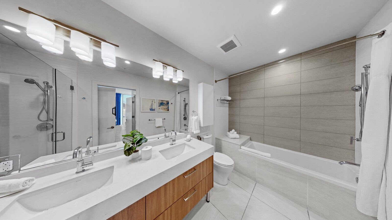 Modern bathroom with a large mirror, double sinks on a wooden vanity, a potted plant, glass shower enclosure, bathtub with beige tile surround, toilet, and bright overhead lighting.