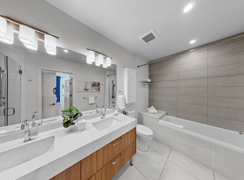 Modern bathroom with double sinks, large mirrors, wooden cabinets, a white toilet, and a bathtub with beige tile. Bright lighting, white walls, and floor tiles create a clean, spacious atmosphere. A small plant decorates the counter.