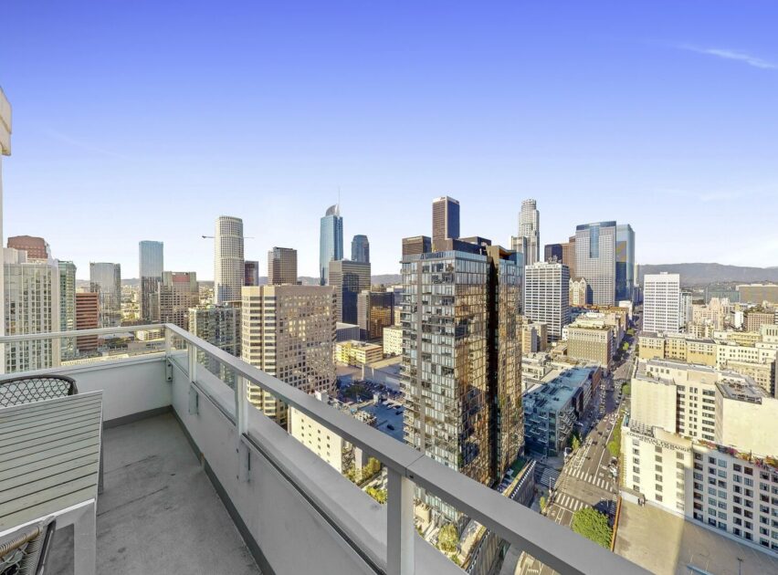 View from a high-rise balcony overlooking a modern city skyline with tall glass buildings under a clear blue sky. The balcony has glass railings and outdoor seating.